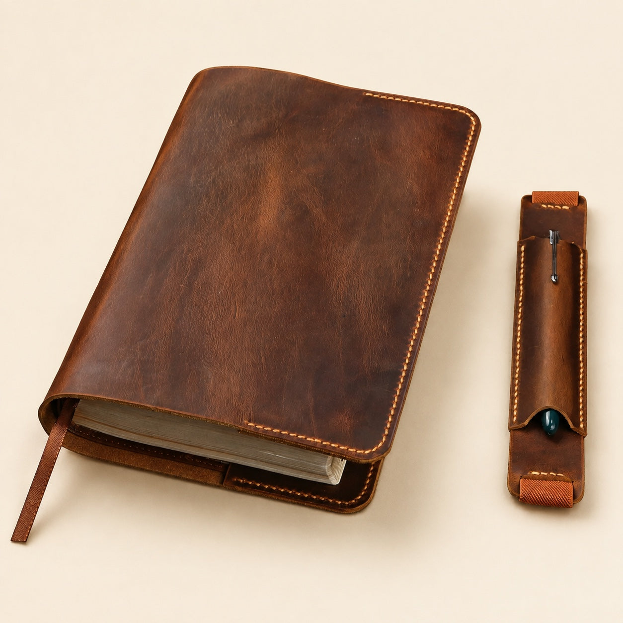 Brown hand-stitched leather notebook cover with matching pen holder, elastic bands and dark green pen on a warm ivory background.
A brown leather planner cover with visible stitching and a pen loop, shown closed with a bookmark ribbon extended, alongside a matching pen in a pen holder, on a warm off-white background.