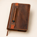 Hand-stitched brown leather notebook cover with integrated pen holder, elastic closures and dark green pen in a premium product setting.
A brown leather journal with a pen loop and pen, photographed on a light beige background.