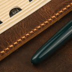 Close-up of a brown leather bible cover  cover featuring thick waxed hand stitching, natural leather grain and a dark green pen.
Close-up of a dark green pen resting on a brown leather surface next to a wooden and leather notebook cover with visible stitching and a partial black logo.